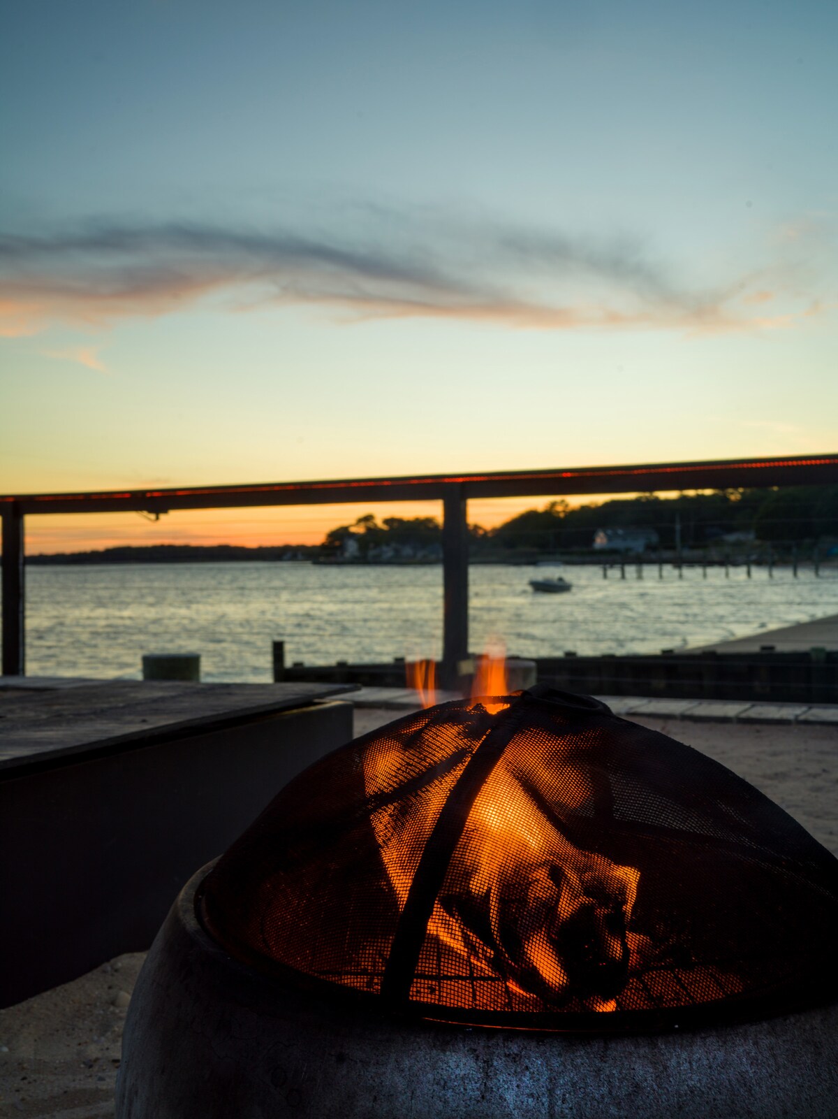 A fire pit is situated on a patio overlooking the water, with warm flames visible through a metal mesh. A serene sunset casts soft colours across the sky, while the bay reflects the fading light, creating a peaceful atmosphere.