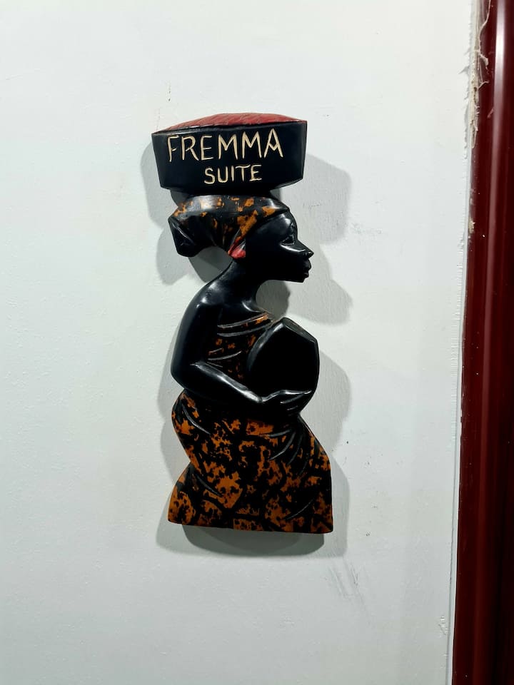 A decorative wall art piece depicting a woman carrying a basket on her head. The artwork showcases rich textures and warm colors, highlighting cultural elements. The sign reads 'Fremma Suite' in bold lettering, adding a welcoming touch to the entrance.