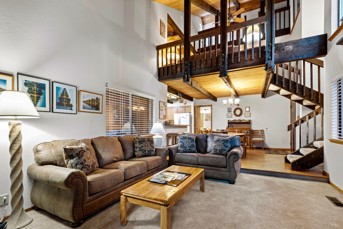 The living room features soft couches arranged around a wooden coffee table. Natural light enters through a window, highlighting framed artwork on the walls. A staircase with wooden railings leads to the upper level, while the dining area is visible in the background.
