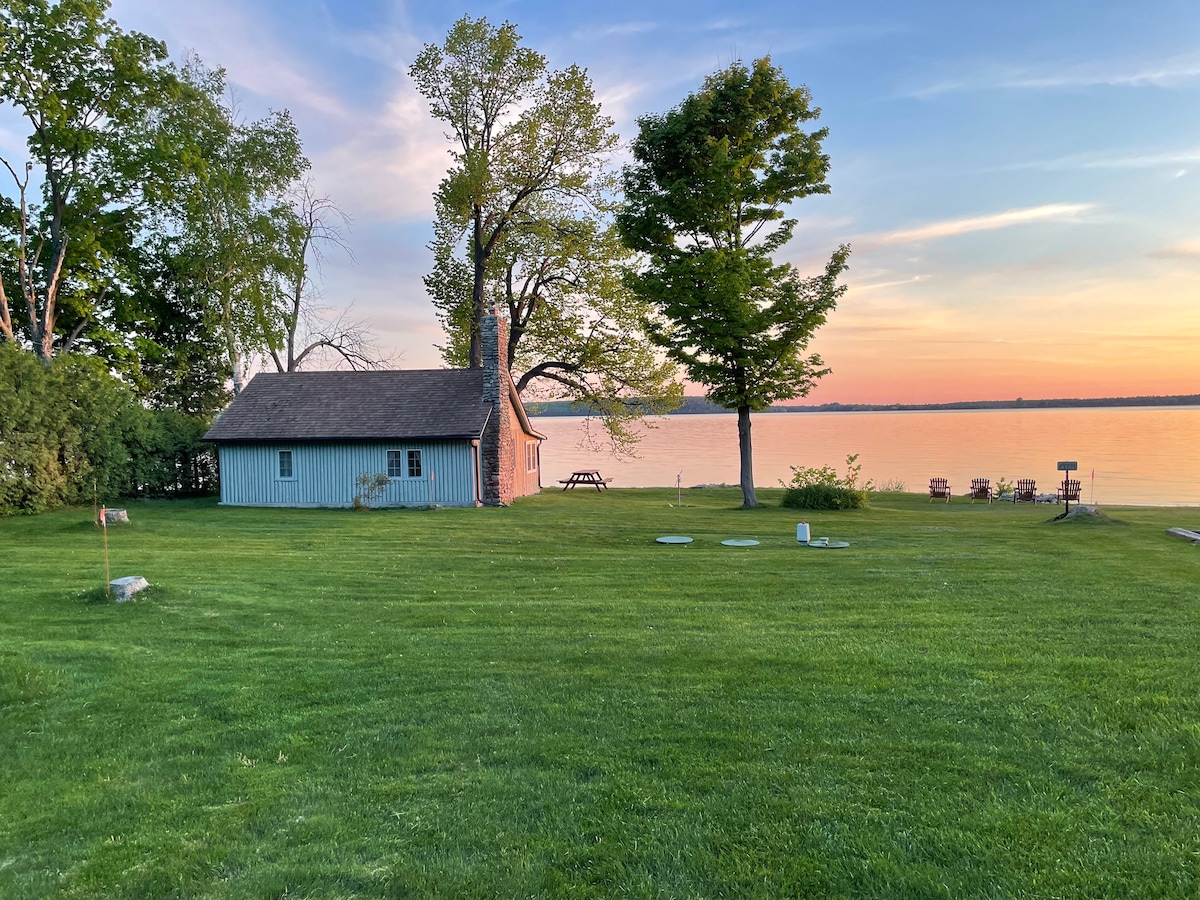 A serene scene captures a rustic cottage nestled among trees, with expansive green grass leading to the bay. The tranquil water reflects the colors of a sunset sky, while several chairs are positioned nearby, inviting relaxation by the water's edge.