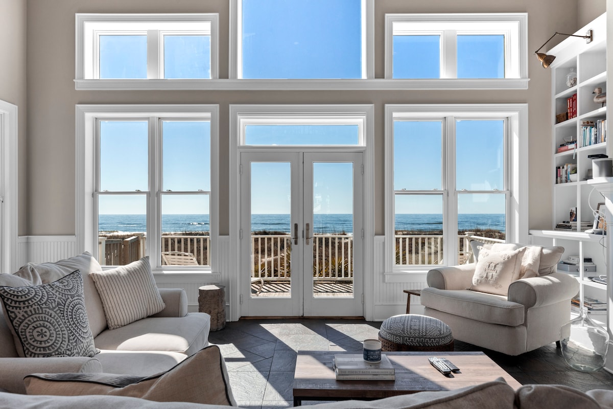 A bright living area is accentuated by large floor-to-ceiling windows, offering uninterrupted views of the ocean. Comfortable seating, including a light-colored armchair and a sofa adorned with neutral pillows, complements the open space, while natural light fills the room.