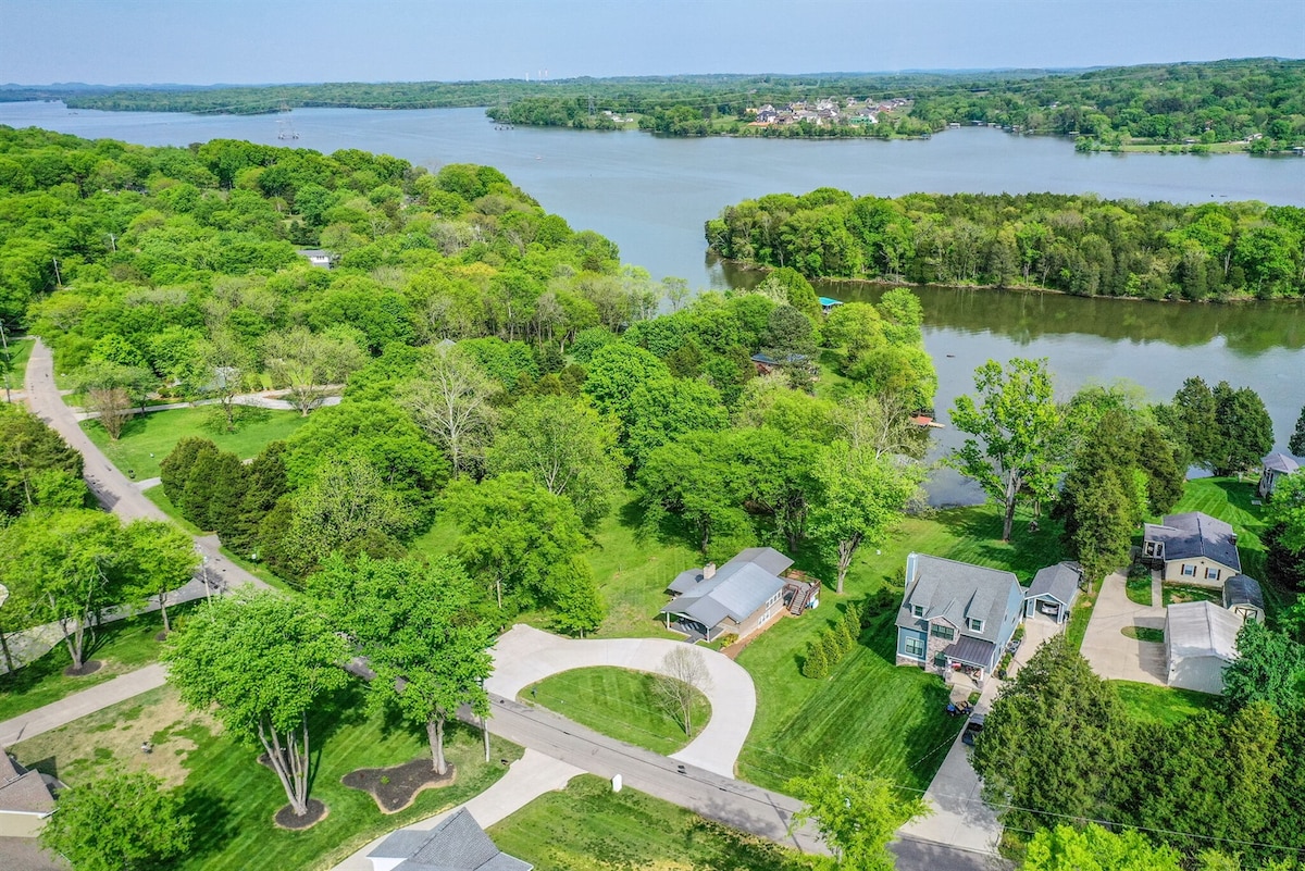 An aerial view captures the lush greenery surrounding the serene Old Hickory Lake. Meandering roads and shaded neighborhoods are visible, showcasing a peaceful lakeside community. The vibrant trees provide a natural frame for the calm water and nearby homes, emphasizing the tranquil environment.