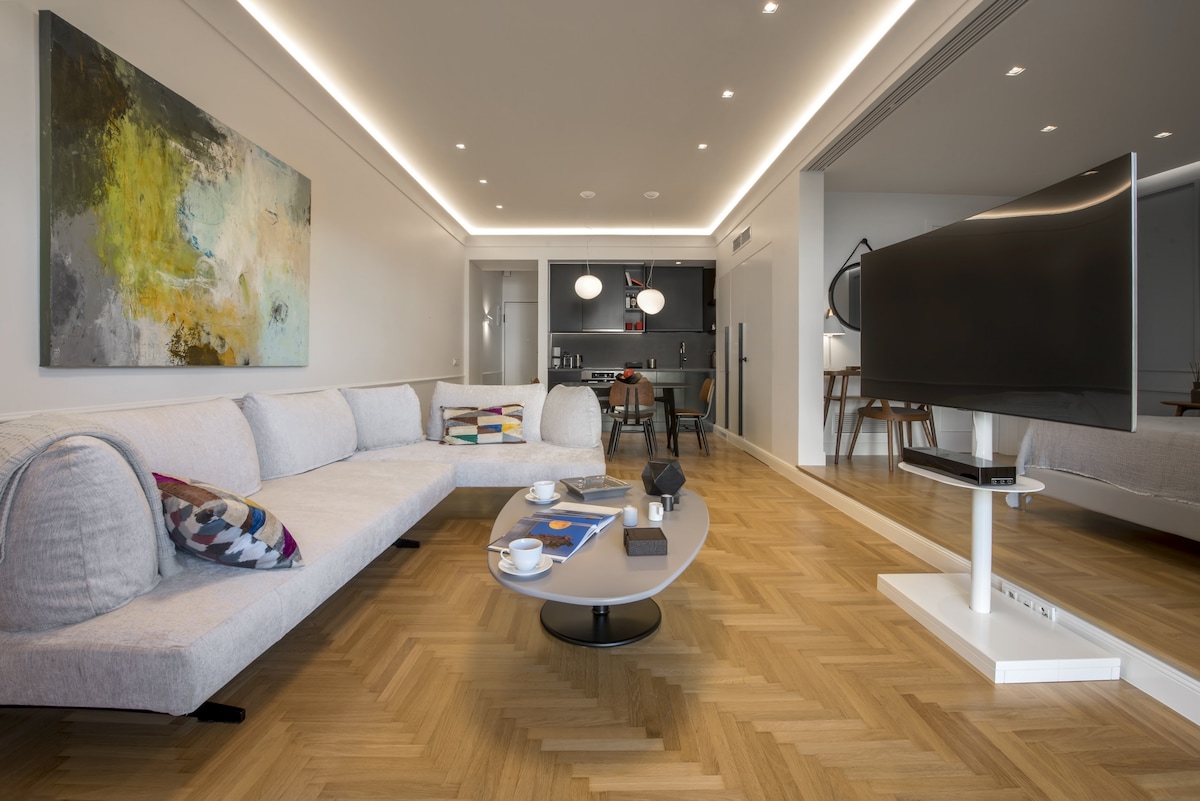 An open-plan living area is showcased with a light-colored sectional sofa adorned with patterned cushions. A low coffee table sits in front, displaying magazines and a decorative object. The space is illuminated by recessed lighting, with a modern television unit visible in the background.