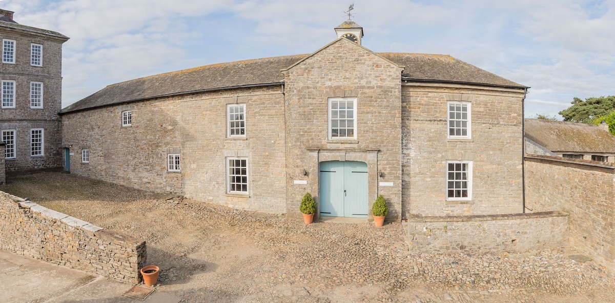 A historic stone building is viewed from the courtyard, showcasing its sturdy construction and large wooden double doors painted in a soft blue. Several windows allow natural light to enter, and potted plants are placed on either side of the entrance, enhancing its welcoming appearance.