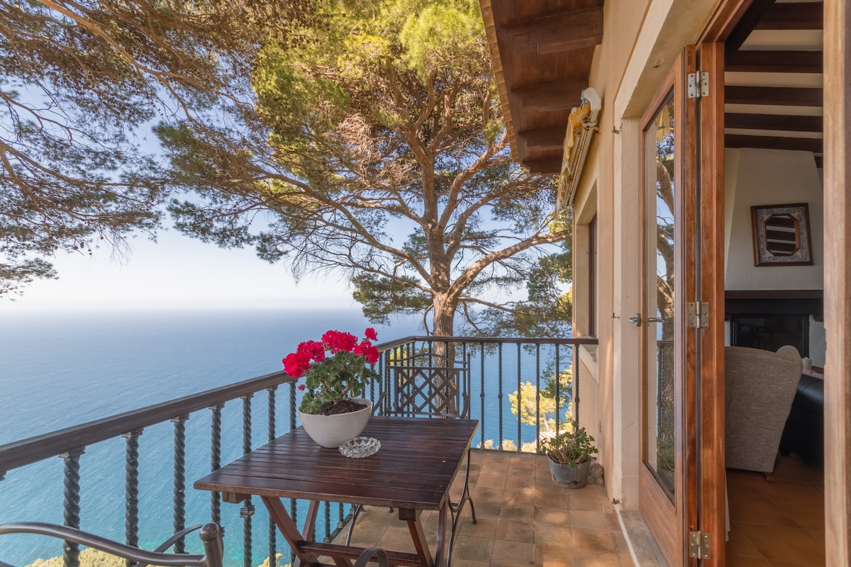 An outdoor terrace features a wooden table set with a decorative bowl, surrounded by blooms in a planter. A railing provides an unobstructed view of the sea and coastline, while sunlight filters through the trees, creating a serene ambiance.
