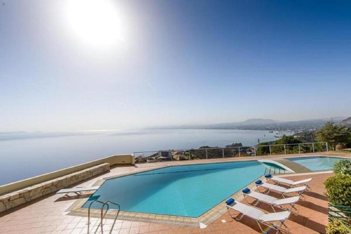 An outdoor pool is shown with a spacious deck and sun loungers arranged around it. The area overlooks a vast sea and distant hills under a clear blue sky, with sunlight reflecting off the water's surface.