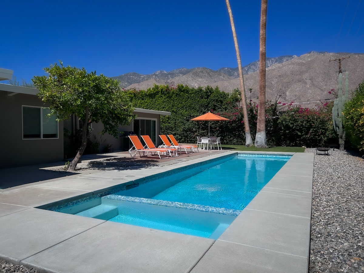 A serene outdoor space features a clear lap pool surrounded by a smooth concrete deck. Lounge chairs with orange cushions are positioned nearby, offering relaxation options. The landscape includes greenery and a mountain backdrop, providing a calm and inviting setting.