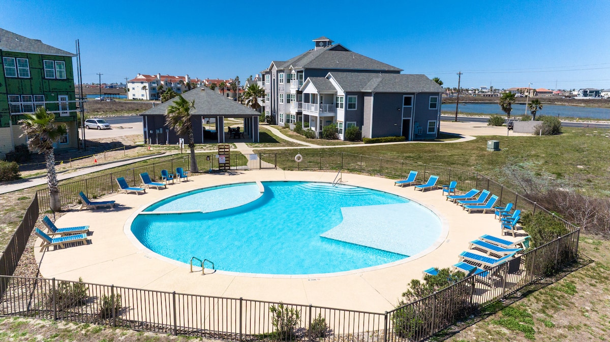A circular heated pool is surrounded by a spacious deck featuring multiple lounge chairs. Lush greenery and palm trees add to the serene setting. The pool area is enclosed by a decorative fence, while distant views of surrounding buildings and open space are visible.