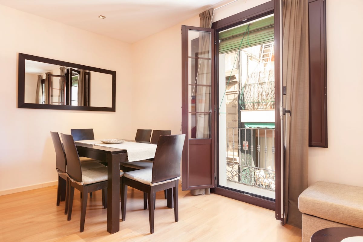 A dining area is depicted with a wooden table surrounded by six dark chairs. A large mirror reflects the space. Floor-to-ceiling glass doors open to a balcony, allowing natural light to fill the room. Light-colored walls and wooden flooring create a warm ambiance.