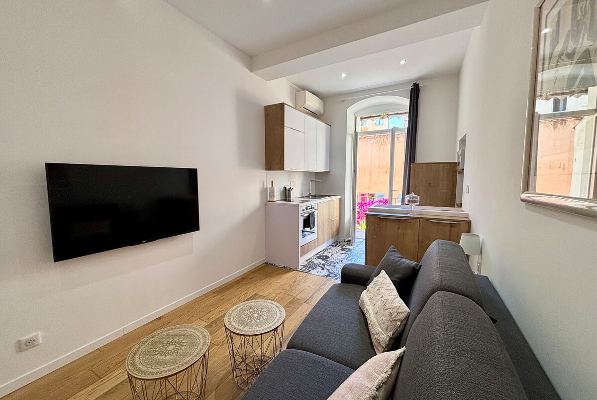 A modern living area features a comfortable gray sofa complemented by two round coffee tables. A wall-mounted television adds functionality, while a kitchenette with wooden cabinetry provides convenience. Natural light enters through a large window, contributing to the welcoming ambiance.
