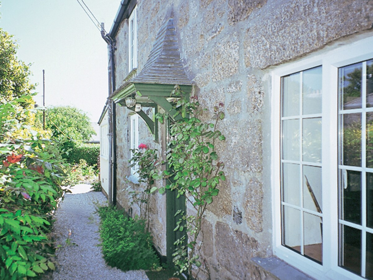The exterior of the 19th-century detached cottage is shown, featuring a stone facade and a charming porch with decorative elements. Lush greenery frames the building, including flowering plants, contributing to a tranquil setting. Bright sunlight illuminates the pathway leading to the entrance.