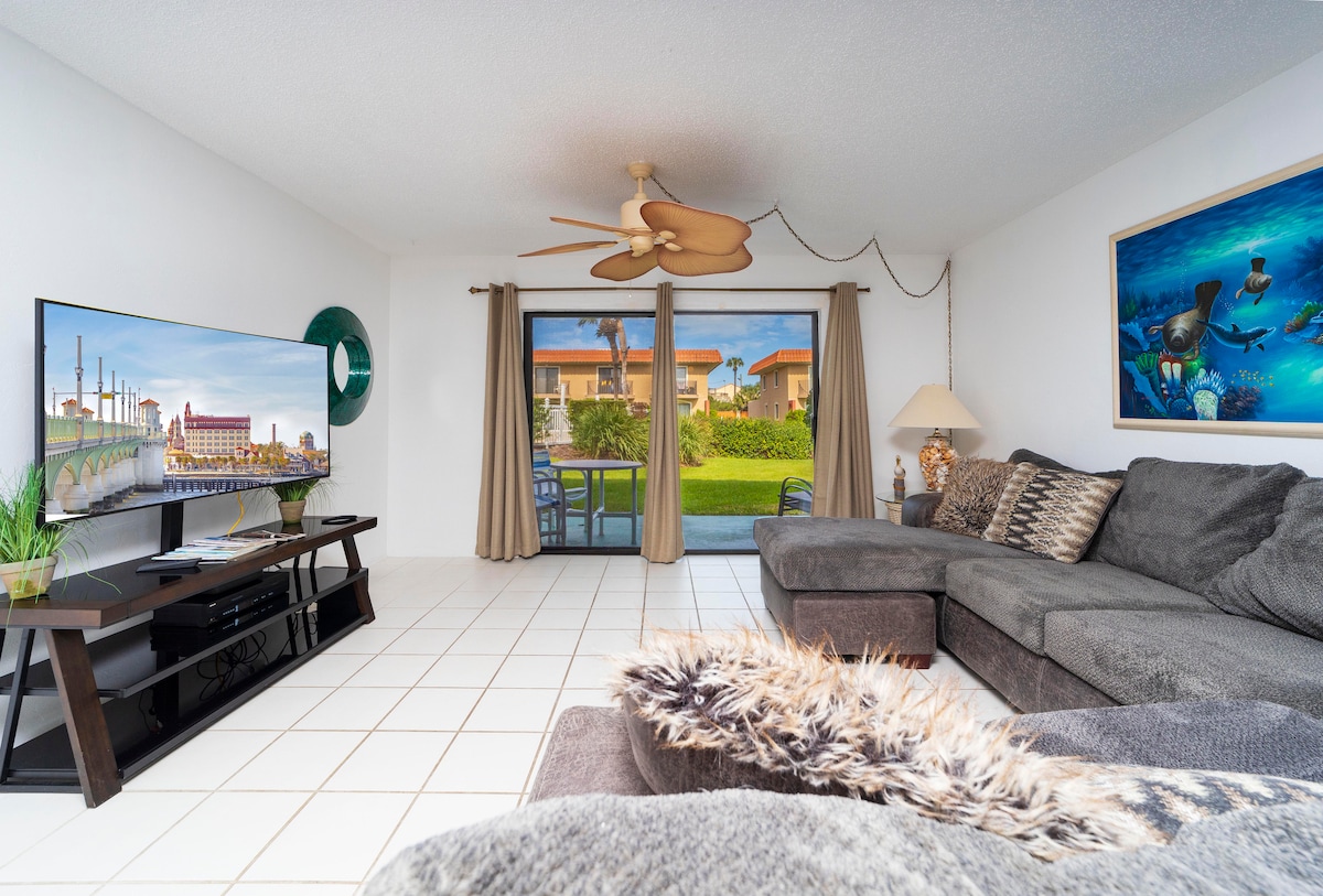 A spacious living area is revealed, featuring a large sectional sofa adorned with decorative pillows. A flat-screen television is mounted on the wall across from the seating. Natural light filters through glass doors that open to a scenic patio with views of the green grounds.