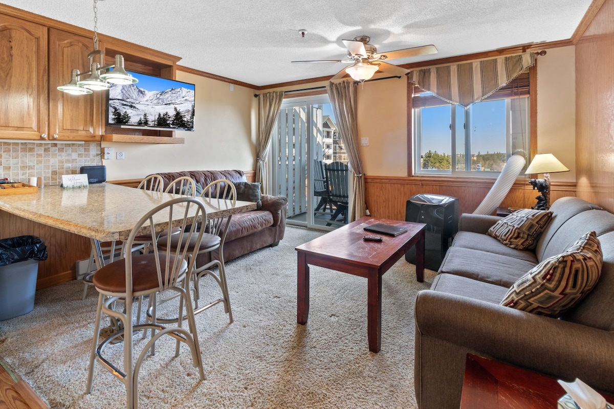 A cozy living area features a comfortable brown leather sofa and a gray fabric loveseat. An open kitchen includes a granite countertop with seating for four. Large windows provide natural light and a view of the balcony outside, while a flat-screen TV is mounted on the wall.