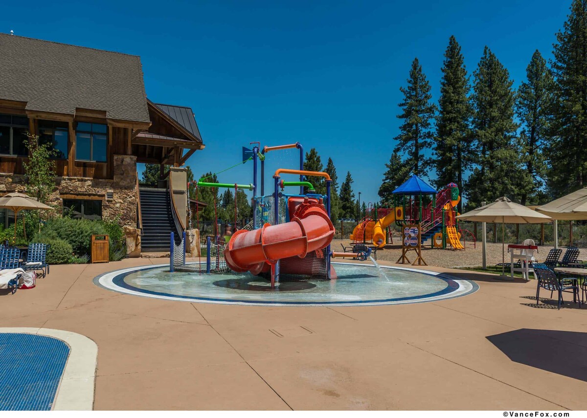 A vibrant water play area is highlighted by a colorful water slide and splash features. Nearby, a playground with climbing structures is visible. Several tables with umbrellas are arranged under the trees, providing shaded seating for guests enjoying the outdoor space.