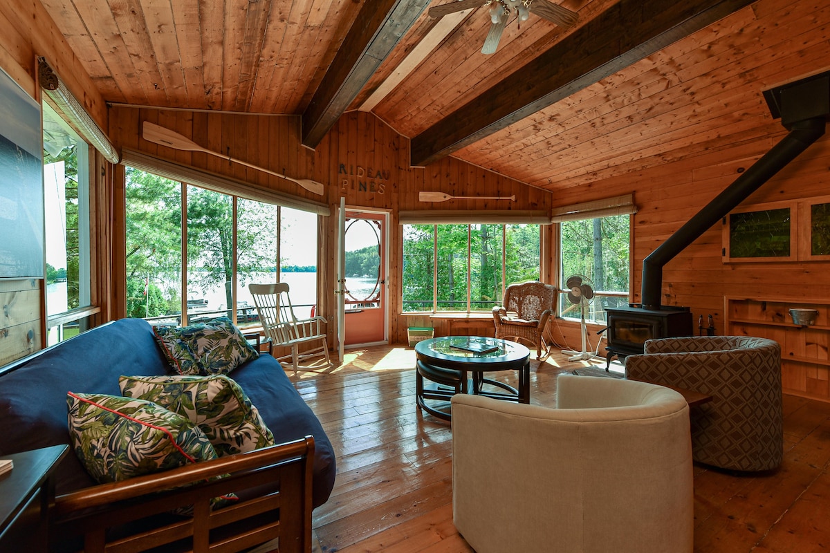 An open-concept living area features large windows framing views of the lake. Natural wood walls create a warm ambiance, while a comfortable seating arrangement includes a blue sofa and a round glass table. A wood stove is visible in one corner, enhancing the cozy environment.