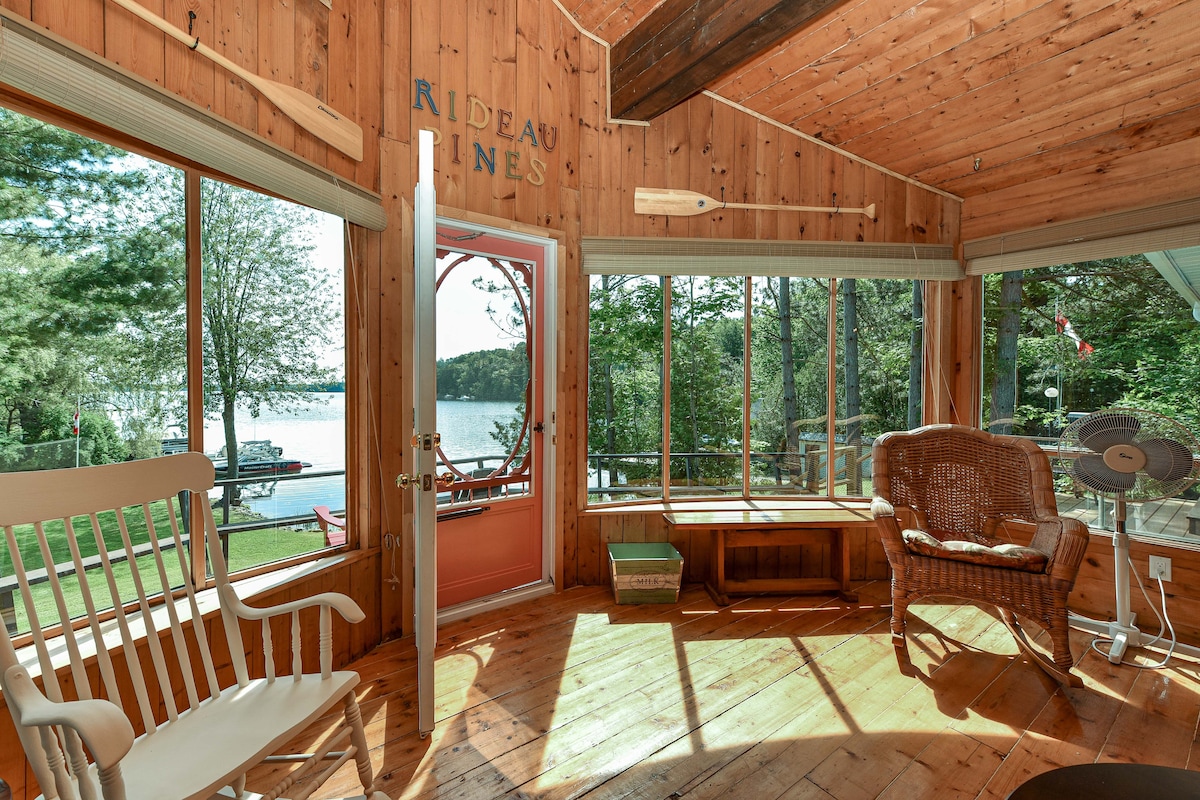 Natural light floods a welcoming space featuring warm wooden walls and flooring. A white rocking chair and wicker chair complement the room. Large windows provide a view of the lake and surrounding greenery, enhancing the connection to the peaceful outdoor environment.