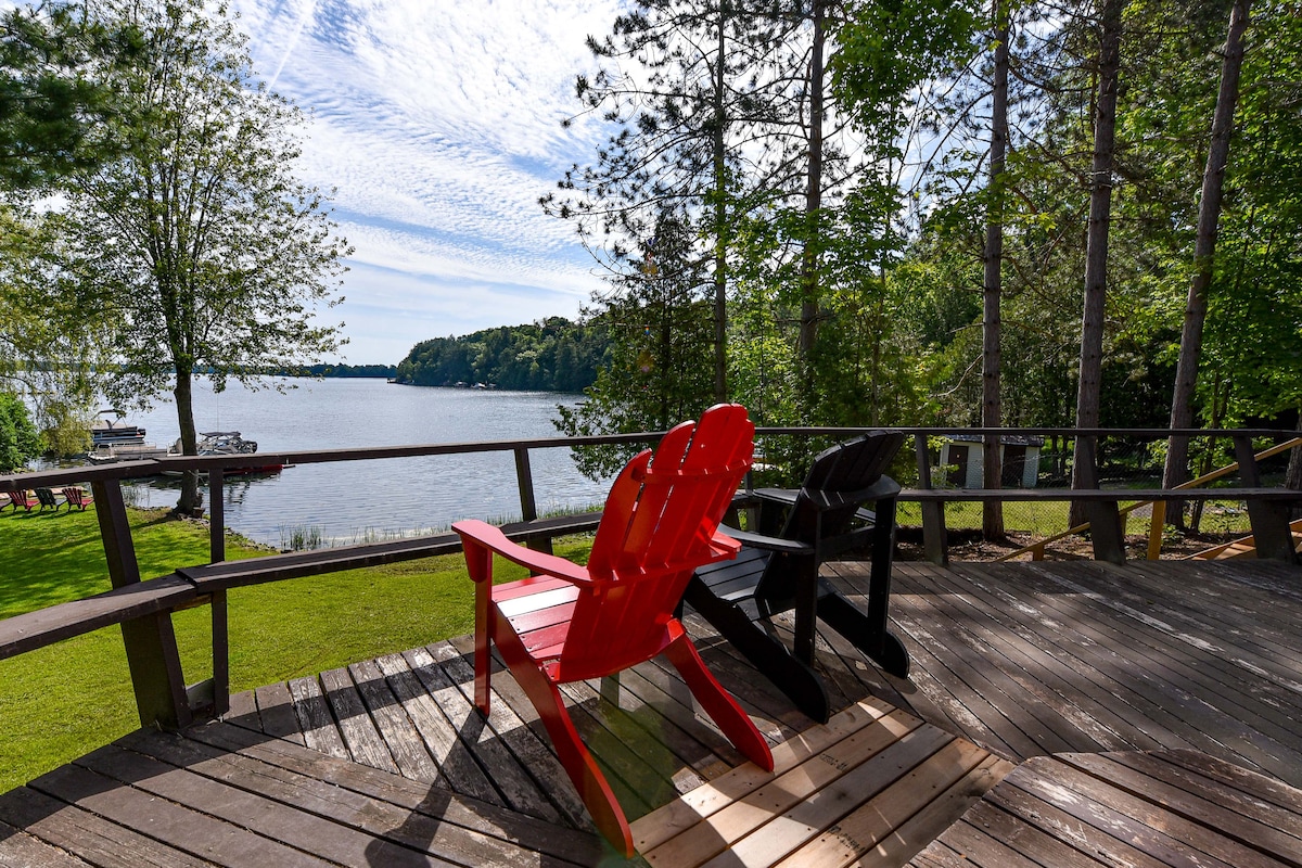 A spacious wooden deck features two Adirondack chairs, one red and one black, positioned to face the serene lake view. Lush greenery surrounds the area, with tall trees providing shade. The calm water reflects a partly cloudy sky, enhancing the peaceful atmosphere.