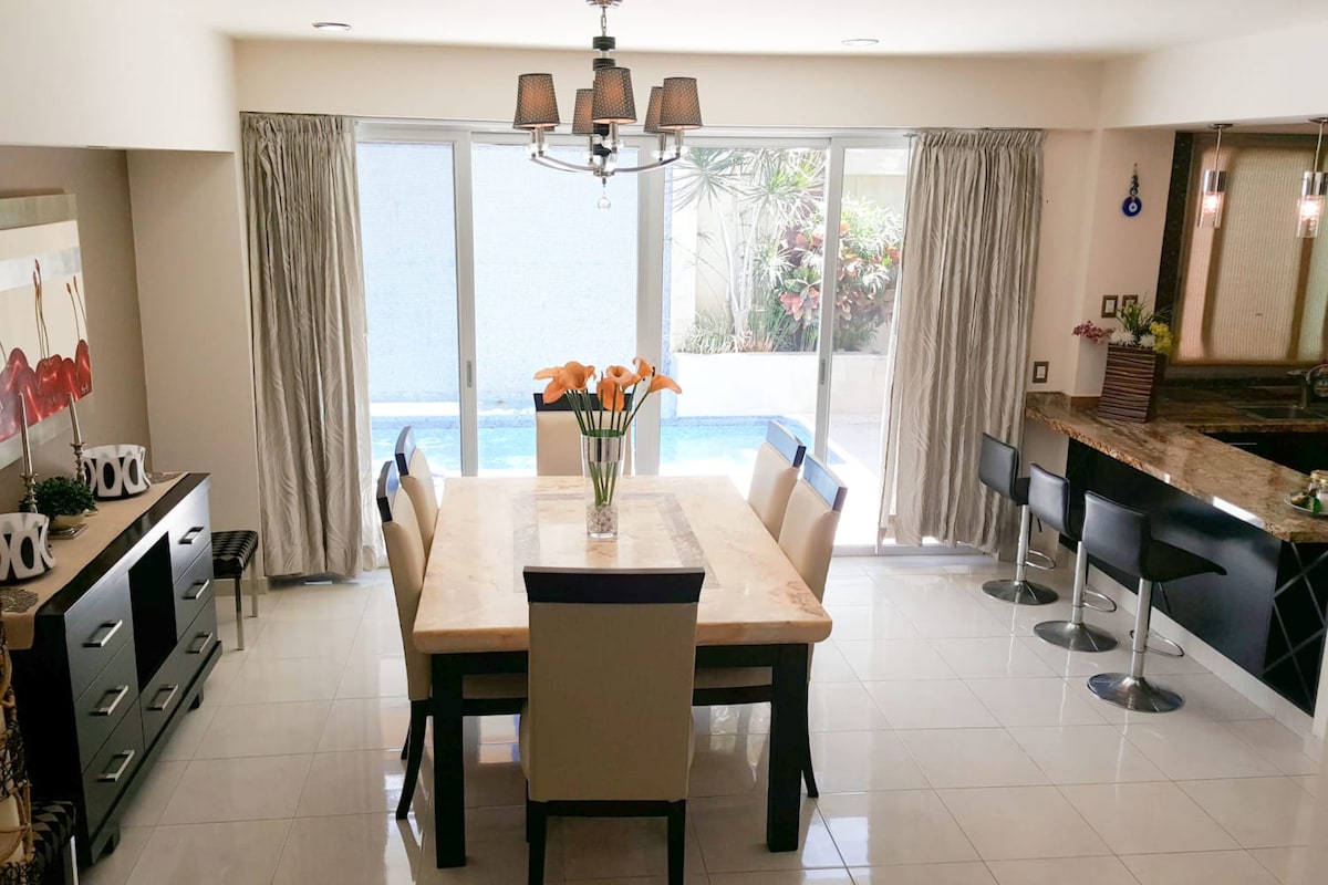 A dining area features a marble table with seating for six, complemented by elegant dining chairs. Large glass doors provide natural light and a view of the pool area, while sheer curtains frame the windows, adding a soft touch to the modern decor.
