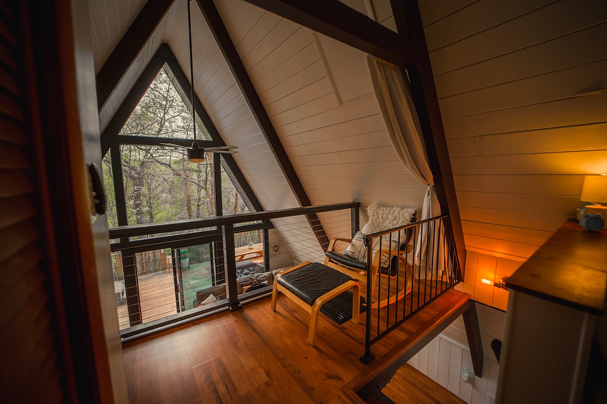 The loft area displays a cozy seating nook with a wooden chair and a lounge chair beside a small side table. Light filters through large glass doors leading to a private balcony with tree views. Wood paneling and warm lighting create a welcoming ambiance.