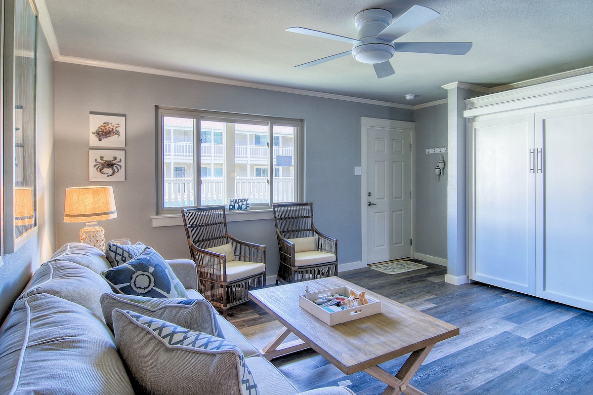 The living area features a comfortable seating arrangement with a soft gray sofa and two wicker chairs. A central coffee table holds a decorative tray. Natural light enters through large windows, illuminating the muted gray walls and enhancing the space's inviting feel.