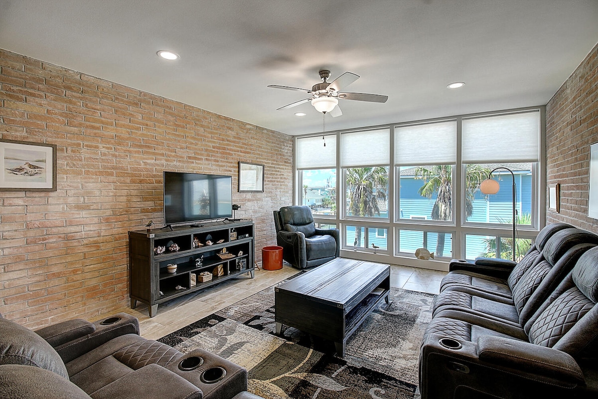 The living area features a mix of textures with a brick wall and cozy seating options, including reclining chairs and a sofa. A coffee table sits in the center, and large windows provide views of the outdoor space. A television is positioned on a media console.