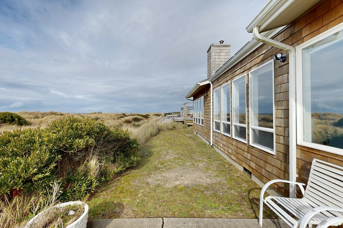 A view from the exterior of the beach house showcases expansive windows along the length of the building. The pathway is bordered by low shrubs and grassy areas, leading towards the ocean. The sky is overcast, creating a calm and muted backdrop.