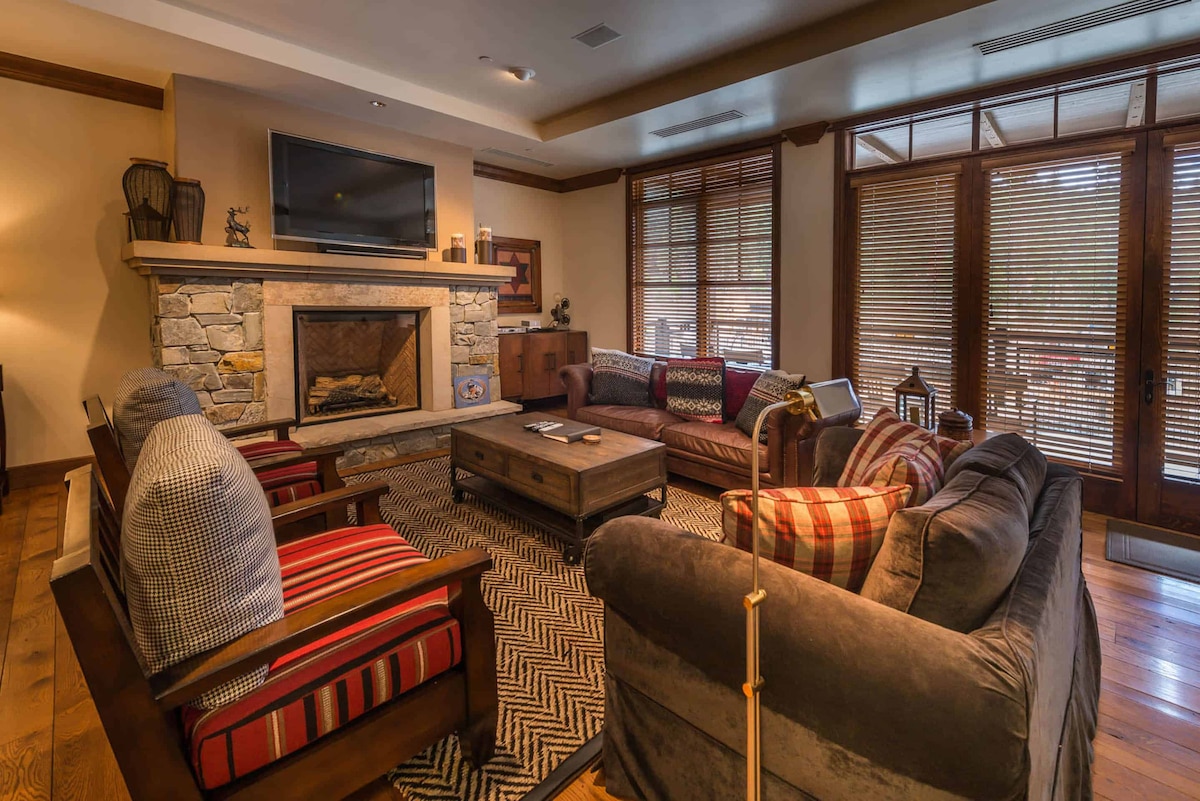 A stylish living area features a stone fireplace as the focal point, surrounded by comfortable seating options. Large windows allow natural light to enter, while patterned rugs enhance the warmth of the space. Dark wood accents create a cozy and inviting environment.