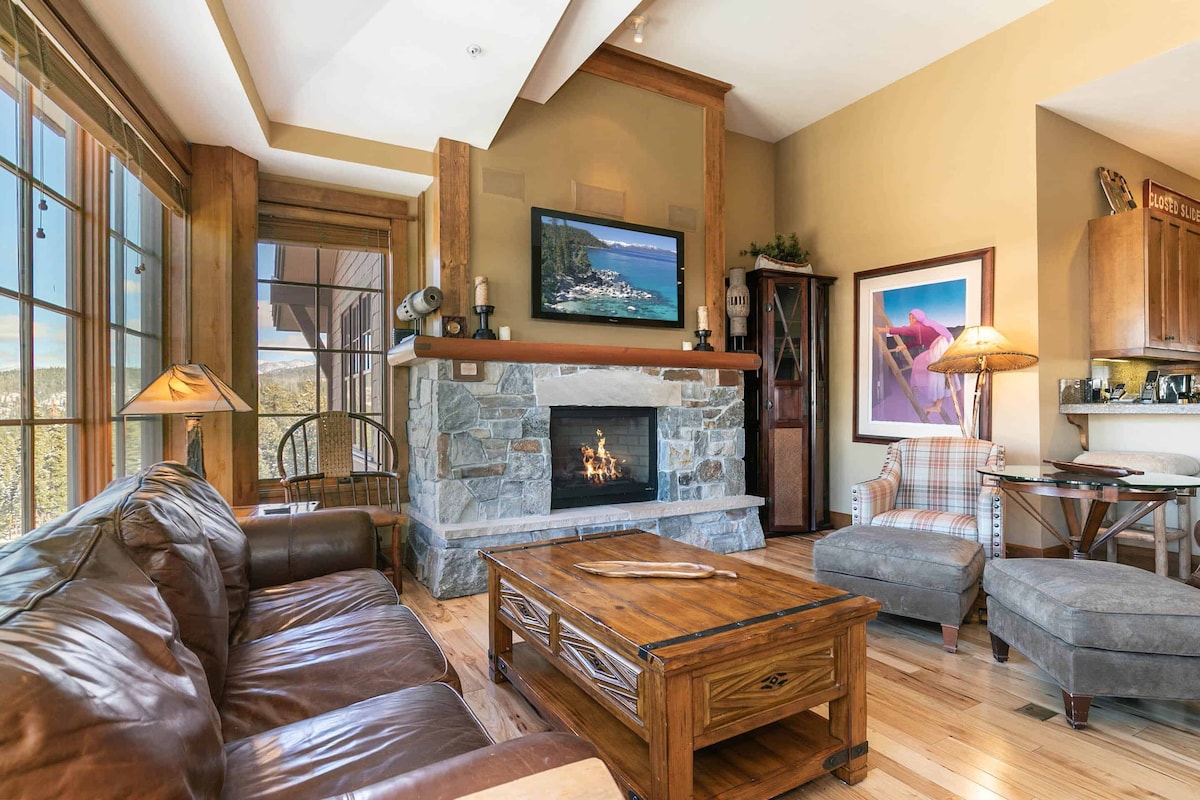 A cozy great room features a stone fireplace as the focal point, surrounded by comfortable seating and wooden accents. Large windows invite natural light and showcase views of the surrounding landscape. A wooden coffee table is centered in the space, complemented by decorative lighting.