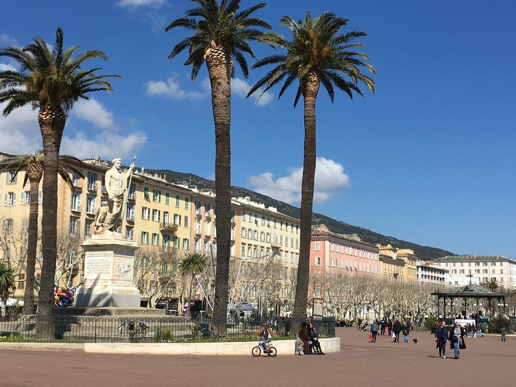 The image features a vibrant public square with iconic palm trees lining the area. A statue stands prominently in the foreground, surrounded by pedestrians enjoying the space. Colorful buildings are visible in the background, under a clear blue sky with scattered clouds.
