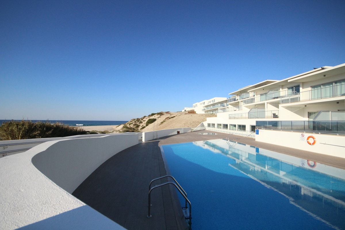 An expansive outdoor pool reflects the clear blue sky, with the ocean visible in the background. The pool area features a sleek, contemporary design with adjacent lounge space. White walls and a paved deck provide a clean, modern aesthetic.