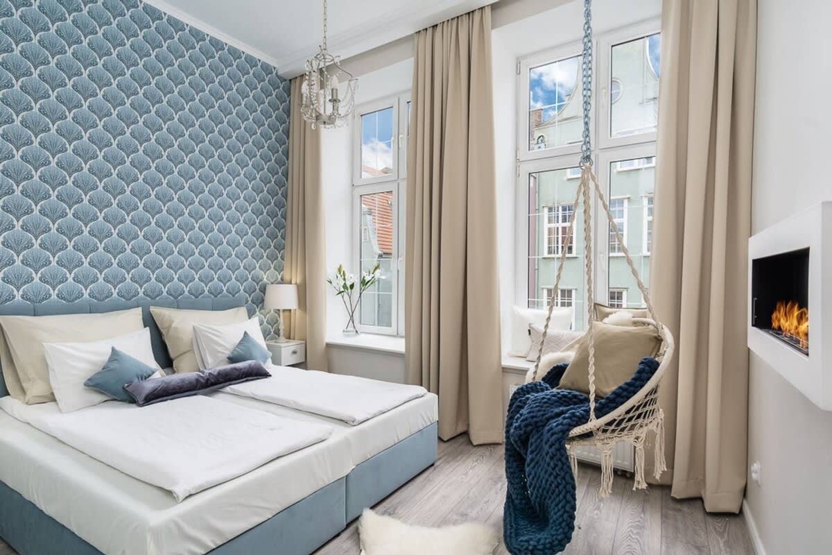 The bedroom is designed with a calming blue accent wall featuring a wavy pattern. A spacious bed with white linens is placed against the wall. A swing chair is positioned nearby, and natural light enters through large windows adorned with beige curtains. A small, modern fireplace is included for added ambiance.