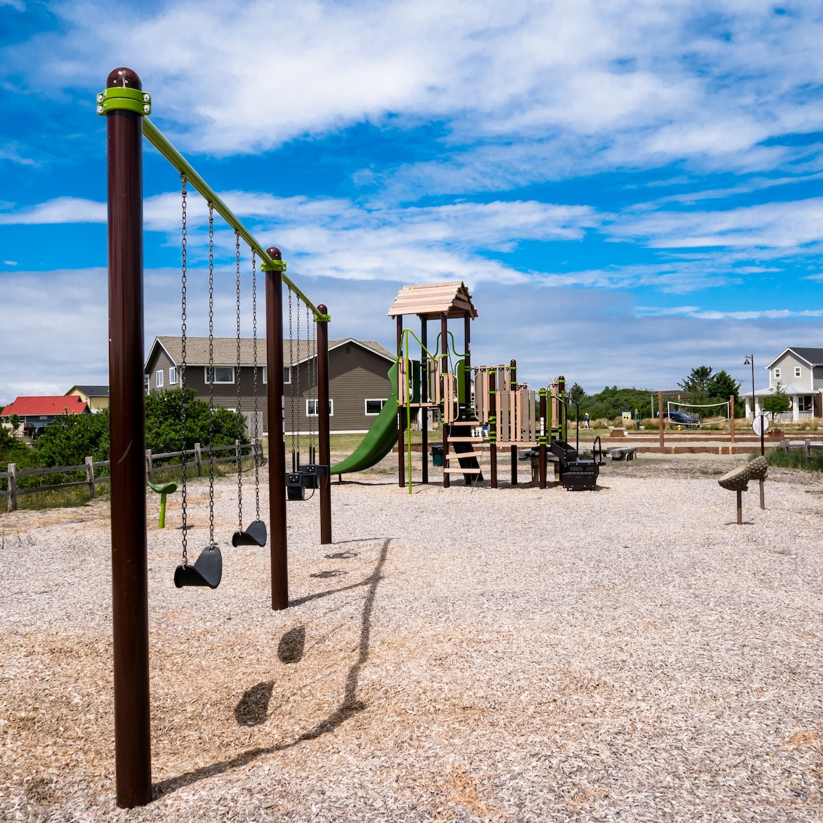 A playground area is showcased, featuring a multi-level play structure with a slide and swings. The ground is covered with soft wood chips, providing a safe play surface. Nearby, homes can be seen against a backdrop of a clear blue sky.