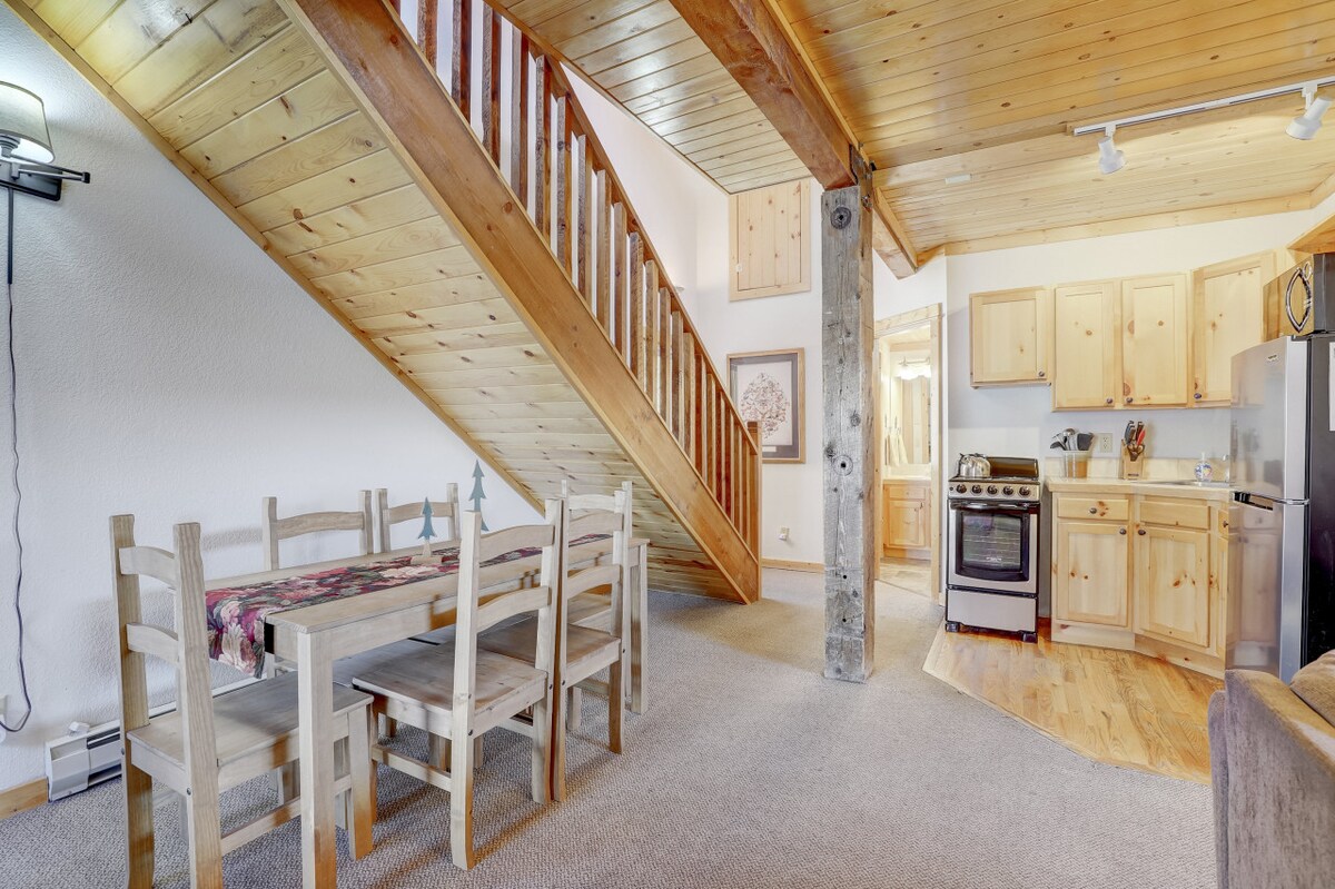 A spacious open-concept area is showcased, featuring a wooden dining table with six chairs beside a staircase leading to the upper level. The kitchen is visible in the background with wooden cabinets and stainless steel appliances. Natural light fills the room through large windows.