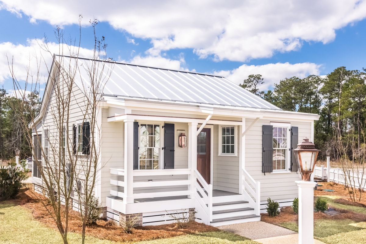 The exterior of Slip Away Cottage features a charming white façade with dark shutters and a metal roof. A welcoming porch with steps leads to the front door, surrounded by landscaped green areas and small trees. Natural light enhances the inviting appearance of the cottage.