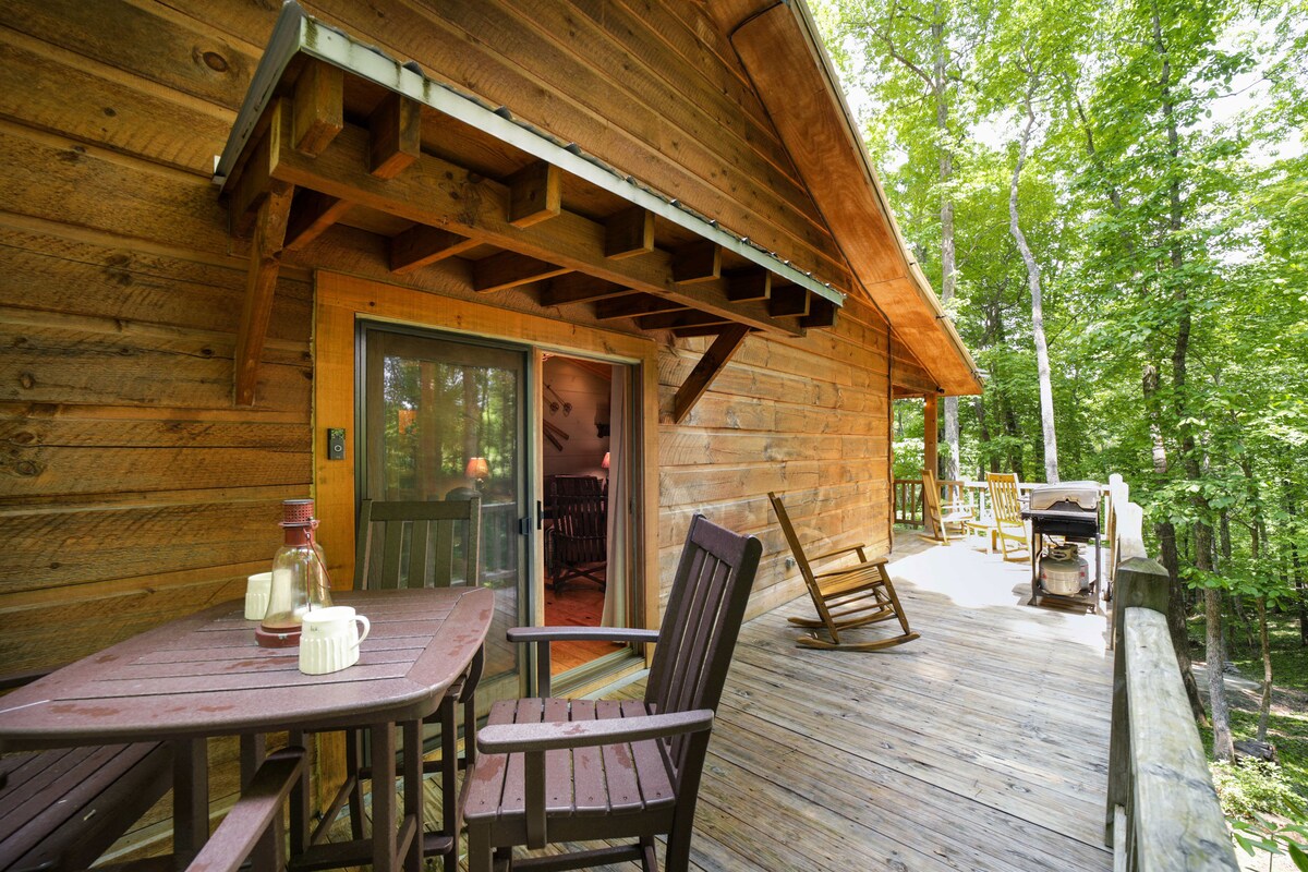 A wraparound deck crafted from natural wood is bordered by lush trees, providing a serene outdoor space. A small dining table with chairs and a rocking chair invite relaxation, while the sliding glass door leads into the cozy interior of the cabin.