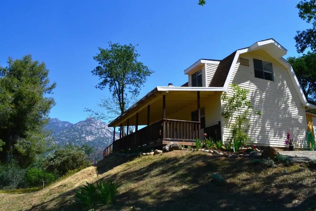 The exterior of a farm-style home is showcased, positioned on a sloping hill under a clear blue sky. The porch extends across the front, inviting relaxation, while trees and colorful flowers enhance the natural surroundings. Mountains are visible in the background, adding to the scenic view.