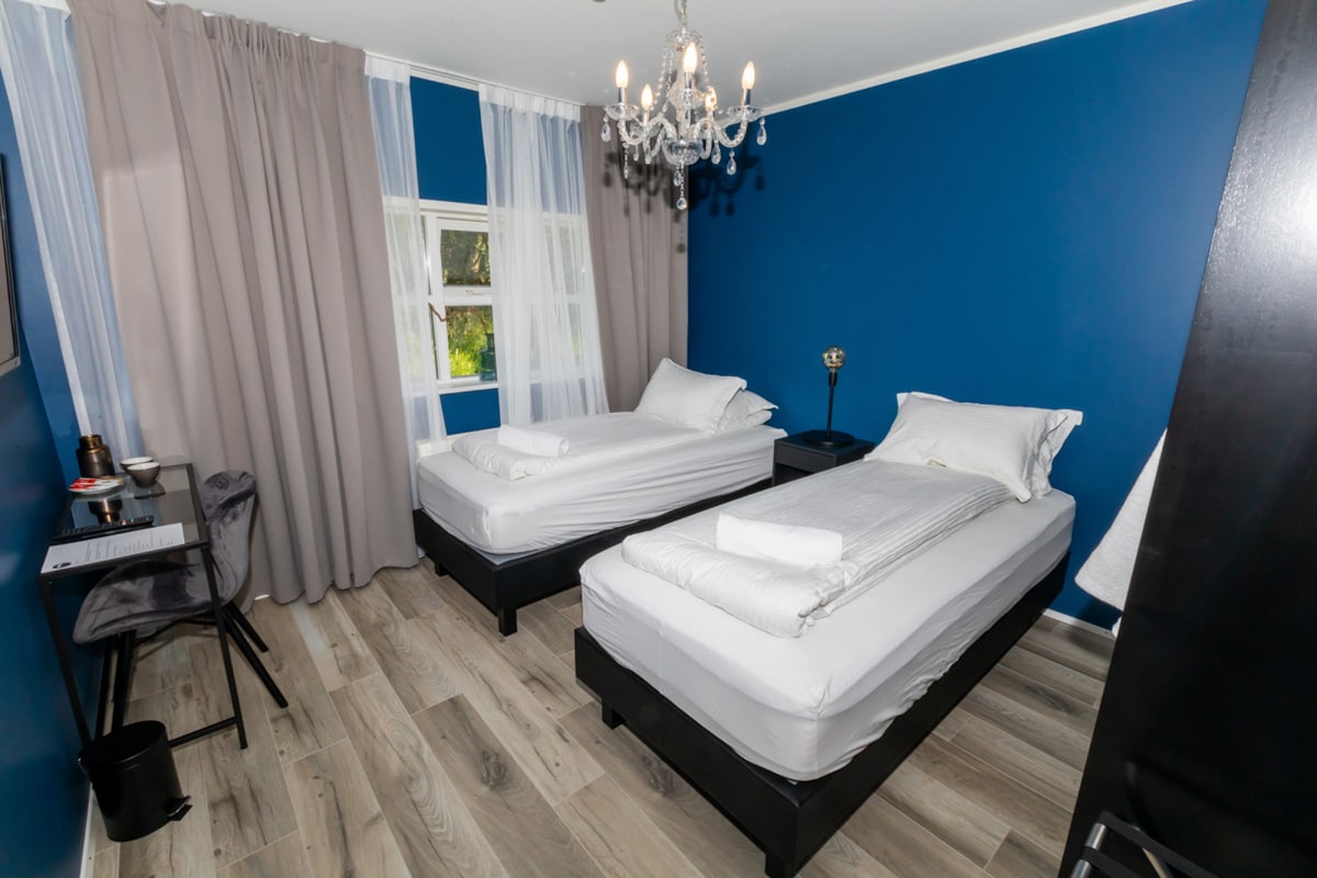 The room features two twin beds with crisp white linens, complemented by a stylish chandelier overhead. A desk and chair are positioned near the window, which is adorned with sheer curtains, allowing natural light to filter in. The accent wall is painted in a calming blue.