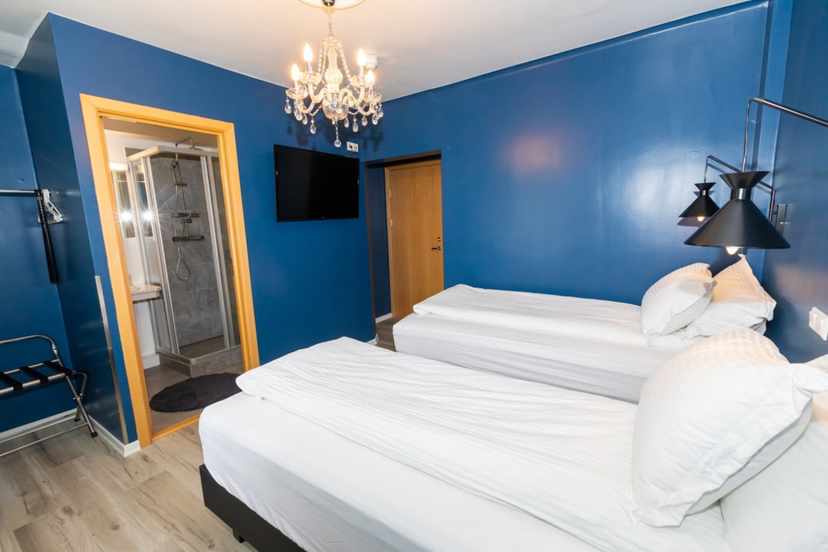 A cozy room is presented with two spacious beds positioned side by side, adorned with crisp white linens. A modern chandelier hangs from the ceiling, complementing the deep blue walls. A private bathroom entrance is visible, along with a TV mounted on the wall.