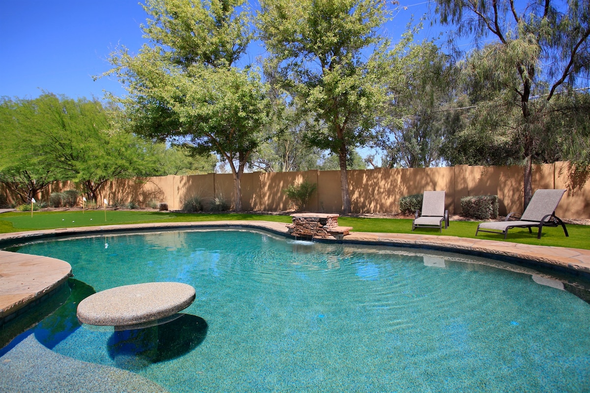 A serene backyard showcases a clear, inviting pool surrounded by verdant grass and neatly trimmed landscaping. Two lounge chairs are positioned nearby, and a circular stone seating area is placed in the shallow end of the pool, inviting relaxation in a private setting.