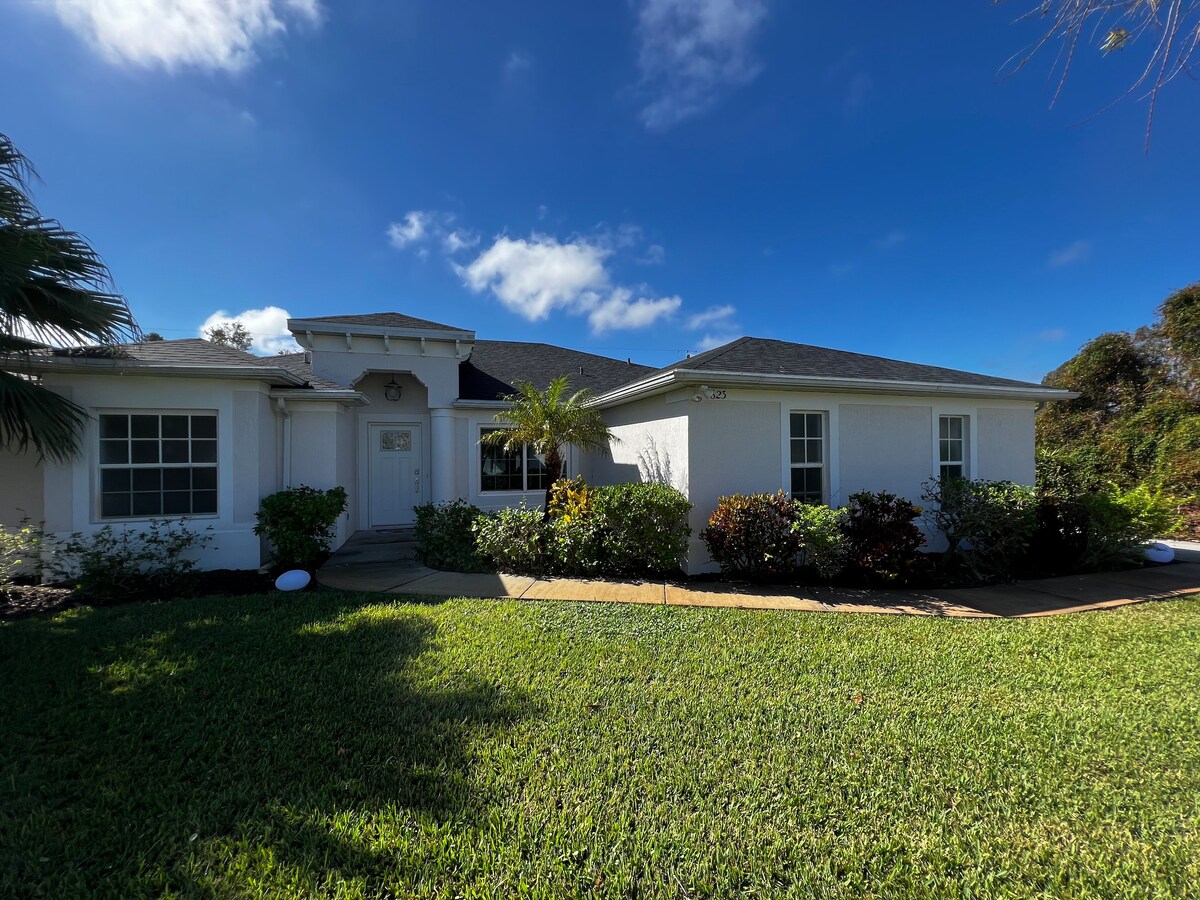 The exterior of a modern villa is displayed, featuring white walls and a welcoming entrance. Lush green grass surrounds the property, while a variety of shrubs and small trees enhance the landscape. Clear blue skies provide a bright backdrop, highlighting the villa's charm.