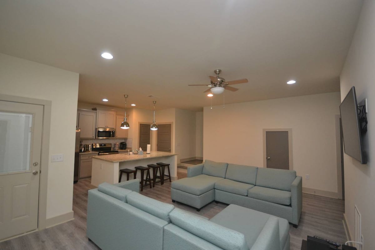 A spacious living area features a light-colored sectional sofa arranged for comfort. Modern pendant lights suspend above a kitchen island with stools. A ceiling fan is installed above, and a flat-screen TV is mounted on the wall, adding to the open-concept layout.
