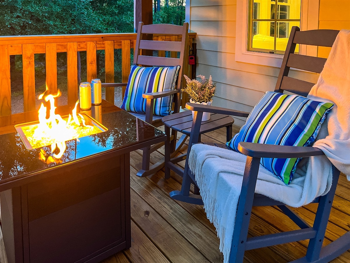 A private deck showcases two wooden rocking chairs adorned with striped cushions. A fire table sits between them, casting a warm glow. A small planter with flowers adds a touch of greenery, while a cozy blanket drapes over one chair, inviting relaxation.