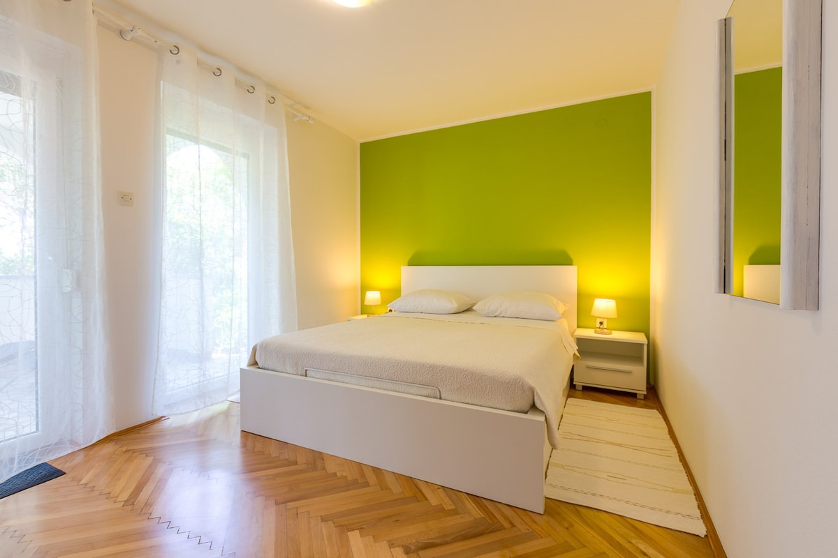 A spacious bedroom features a large bed with light bedding, complemented by modern bedside tables and lamps. A vibrant green accent wall adds color, while natural light filters in through sheer curtains. A mirror reflects the room's bright atmosphere, enhancing the overall open feel.