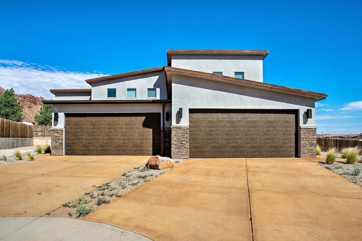 The exterior of a modern two-story townhome is depicted, featuring a combination of stone and stucco facades. The spacious driveway leads to three garage doors, providing ample parking space. Clear blue skies complement the structure, while desert landscape elements are visible in the foreground.