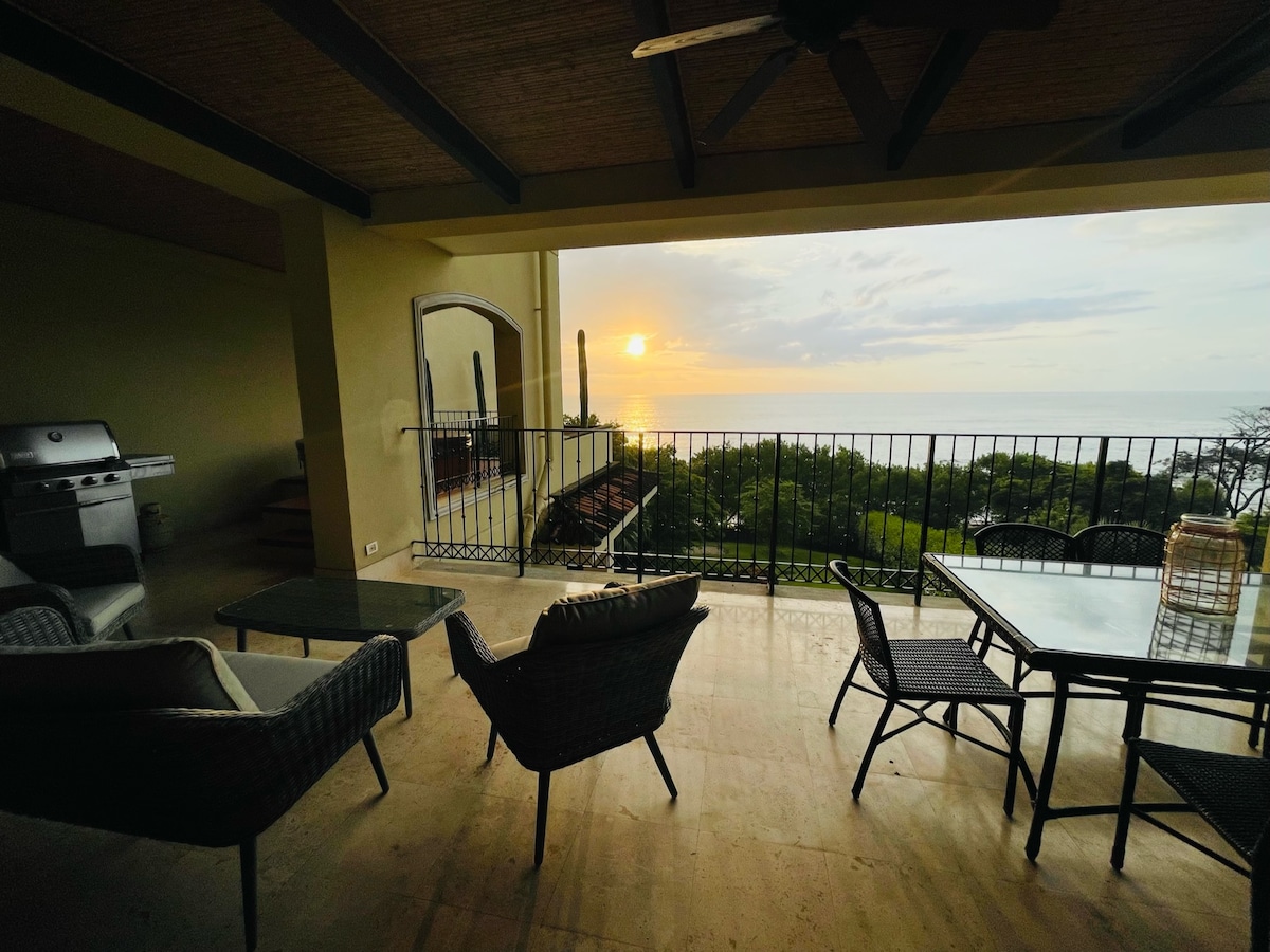 A shaded outdoor terrace features comfortable seating arrangements, including two cozy chairs and a glass-top table. The expansive view reveals a sunset over the ocean, with vibrant hues reflecting on the water, and lush greenery in the foreground.