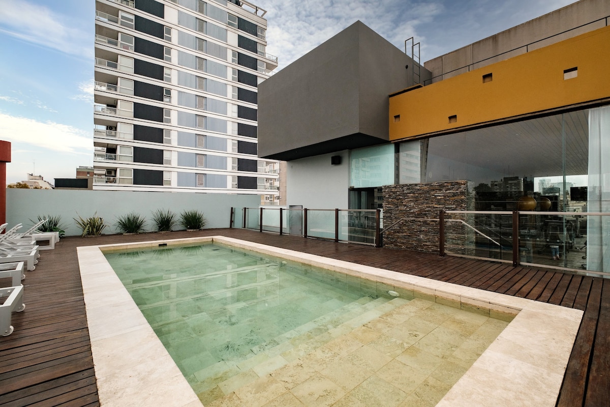 A spacious rooftop terrace features a swimming pool surrounded by a wooden deck. Lounge chairs are arranged along one side, with planters adding greenery to the area. The backdrop includes a modern building, showcasing the urban landscape of the city.