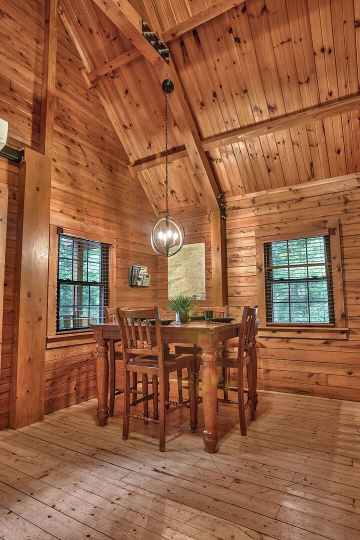 A wooden dining area features a square table surrounded by four classic chairs. The space is illuminated by a round pendant light hanging from a high ceiling, while natural light filters through two windows adorned with wooden blinds.