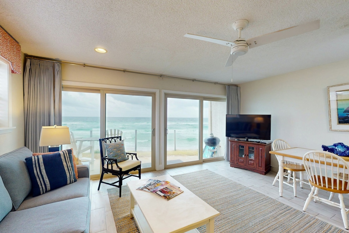 The living area features large glass doors that open to the beachfront, allowing natural light to fill the space. A comfortable sofa and chair are arranged around a coffee table, with a TV positioned on a wooden stand. A dining table with chairs is present near the windows, enhancing the inviting setting.