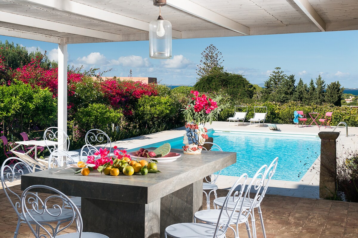 An outdoor dining area is presented beneath a shaded pergola, featuring a stone table adorned with a colorful display of fruits. The scene is complemented by a private swimming pool visible in the background, surrounded by vibrant gardens and lounge chairs.