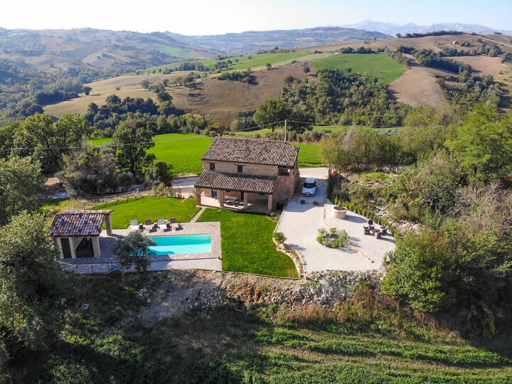 An aerial view of a restored farmhouse is presented, surrounded by lush green hills and farmland. The property features a private pool, a spacious garden, and a covered outdoor relaxation area. The picturesque landscape highlights the tranquil countryside ambiance.